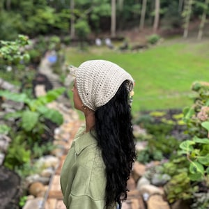 May include: A woman wearing a light brown crocheted bandana with a green jacket and blue jeans. The bandana is tied around her head and covers her forehead. She has long, dark hair.