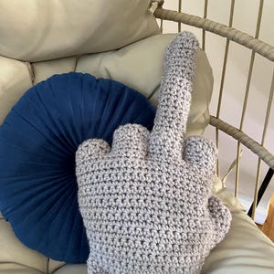 May include: A crocheted pillow shaped like a middle finger, in a light gray color, sitting on a white couch with a blue round pillow behind it.