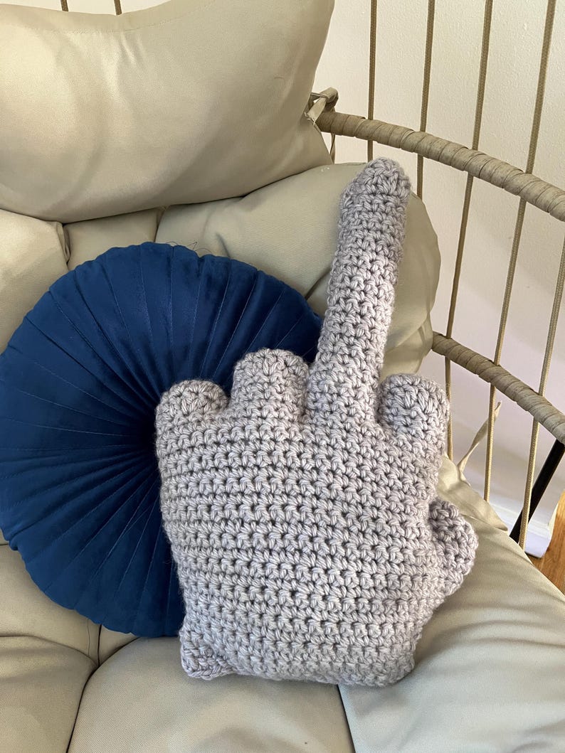 May include: A crocheted pillow shaped like a middle finger, in a light gray color, sitting on a white couch with a blue round pillow behind it.