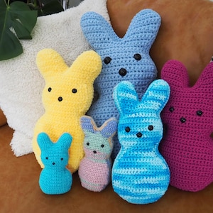 May include: A collection of six handmade crochet bunny plushies in various sizes and colors. The bunnies are in shades of yellow, blue, pink, and a blue and white striped pattern. Each bunny has black button eyes and a cute, rounded shape.