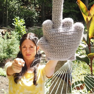 May include: A woman is holding up a large crocheted gray middle finger. She is wearing a yellow shirt and has her hair pulled back. The finger is in the foreground and the woman is in the background.