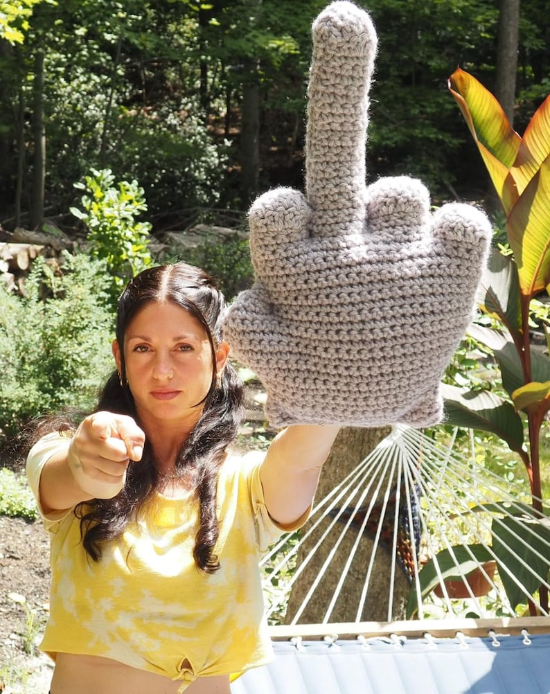 May include: A woman is holding up a large crocheted gray middle finger. She is wearing a yellow shirt and has her hair pulled back. The finger is in the foreground and the woman is in the background.