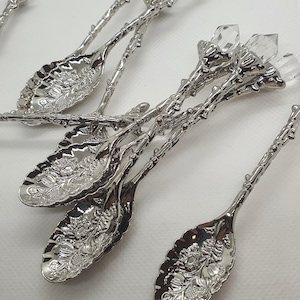 Puede incluir: Conjunto de cucharas plateadas ornamentadas. Cada cuchara presenta un diseño floral detallado en el cuenco y un adorno de cristal en el extremo del mango. Las cucharas están dispuestas sobre una superficie blanca.