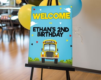 May include: Welcome to Ethan's 2nd Birthday sign with a yellow school bus illustration on a blue background. The sign is on an easel with yellow, black, and blue balloons in the background.