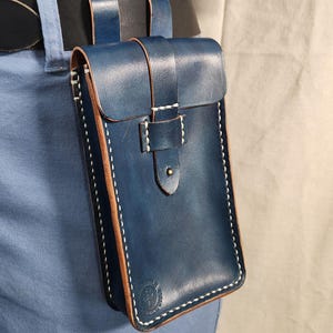 May include: A blue leather belt pouch with a flap closure, attached to a black belt. The pouch has white stitching and a brown edge. The pouch is designed to be worn on the hip. The overall theme is leather goods.