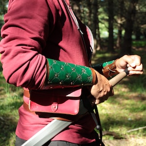 May include: Green and brown leather arm guards with a diamond pattern and gold studs. The arm guards are worn over a burgundy long-sleeve shirt. A burgundy leather pouch and belt are also visible.