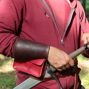 May include: A brown leather arm guard and a red leather pouch with a buckle are visible. The arm guard is worn on the forearm. The pouch is attached to a brown leather belt. A sword is partially visible.