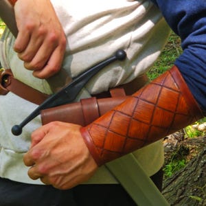 May include: Close-up of a brown leather arm guard with a diamond pattern. The arm guard is worn over a blue sleeve. A black sword and brown leather sheath are visible, along with a white shirt and brown belt.