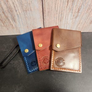 May include: Three leather cardholders in blue, red-brown, and brown, each with a flap closure and a brass snap. The cardholders are stitched with contrasting thread and feature a stamped logo that reads "CALIBURN LEATHERWOOD".