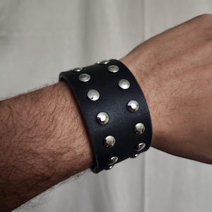 May include: A black leather cuff bracelet with silver-colored studs. The wide bracelet is worn on a person's wrist. The studs are evenly spaced across the surface of the bracelet. The bracelet is a bold accessory.