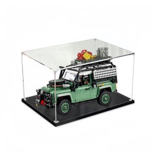 Może przedstawiać: Zielony model Lego Land Rover Defender z czarnym bagażnikiem dachowym i białym dachem, prezentowany w przezroczystej akrylowej gablocie z czarną podstawą.