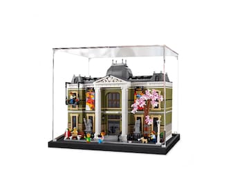 Può includere: Un modello LEGO verde di un museo d'arte con una cupola bianca, un albero di ciliegio in fiore e un cartello che recita "The Museum". Il modello è esposto in una teca di acrilico trasparente con una base nera.