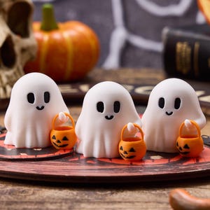 Könnte beinhalten: Drei weiße Geisterfiguren mit schwarzen Augen und Lächeln, die jeweils einen orangefarbenen Kürbis-Eimer halten. Die Geister befinden sich auf einem dekorativen Halloween-Teller, mit einem Schädel und einem Kürbis im Hintergrund.