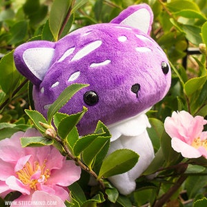 May include: A purple mushroom plush toy with white spots and cat ears, set amongst green foliage and pink flowers. The toy has black eyes and a white collar. The website address www.stitchmind.com is visible.