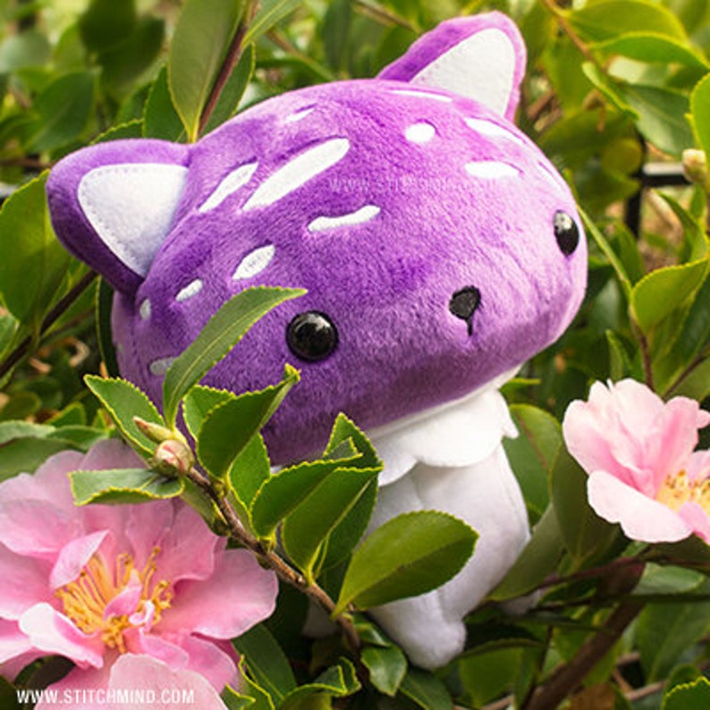 May include: A purple mushroom plush toy with white spots and cat ears, set amongst green foliage and pink flowers. The toy has black eyes and a white collar. The website address www.stitchmind.com is visible.