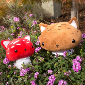 May include: Two plush mushroom-shaped toys with cat-like features sit amongst purple flowers. One is red with white spots, the other is brown. Both have black eyes and small noses. The toys are surrounded by green foliage.