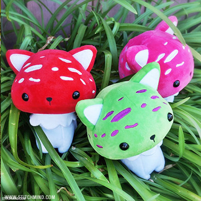 May include: Three plush mushroom toys in red, green, and pink, each with white spots and cat-like ears. The toys have black eyes and white undersides, set against a backdrop of green foliage.