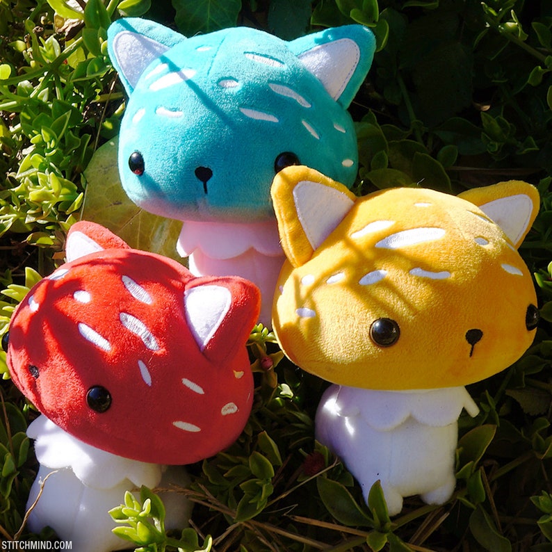 May include: Three plush mushroom toys in red, yellow, and teal, each with white accents and a cat-like face. The toys have white stems and are set against a background of green foliage.