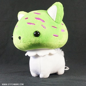 May include: A soft toy with a green head, decorated with pink markings and large black eyes. The ears have white inner details. The body is white with a ruffled collar. A small, white tail is visible. A whimsical, handmade item.
