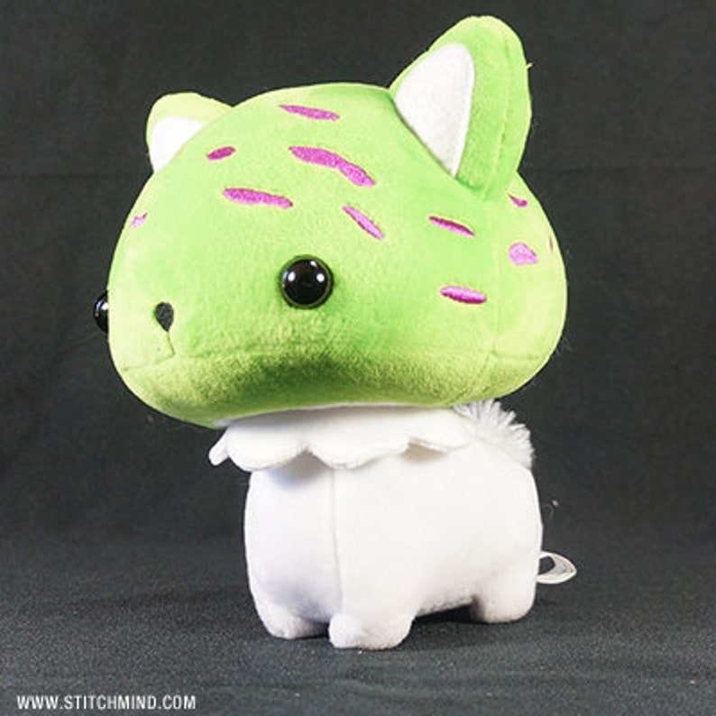 May include: A soft toy with a green head, decorated with pink markings and large black eyes. The ears have white inner details. The body is white with a ruffled collar. A small, white tail is visible. A whimsical, handmade item.