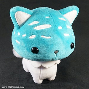 May include: A soft toy cat with a blue head and white spots, white ears, and a white body. The cat has large black eyes and a small, embroidered nose. The toy is made of soft fabric and has a cute, whimsical design.