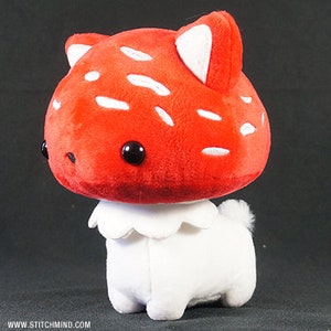 May include: A soft toy shaped like a mushroom-dog hybrid. The red mushroom cap has white spots and black eyes. The white body has a frilly collar and a small tail. The toy is designed to look like a cute, cartoonish creature.