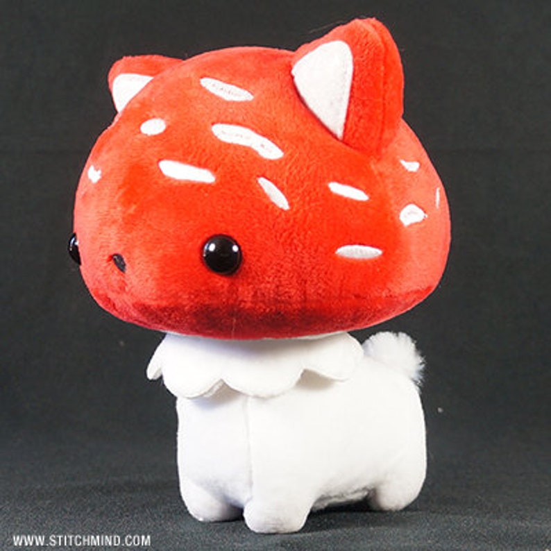 May include: A soft toy shaped like a mushroom-dog hybrid. The red mushroom cap has white spots and black eyes. The white body has a frilly collar and a small tail. The toy is designed to look like a cute, cartoonish creature.