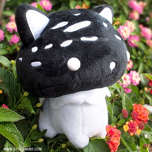May include: A black mushroom-shaped plush toy with white spots and cat ears, set against a backdrop of flowers. The toy has a white base. The website address "www.stitchmind.com" is visible at the bottom.