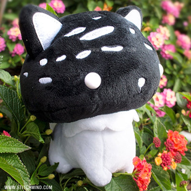 May include: A black mushroom-shaped plush toy with white spots and cat ears, set against a backdrop of flowers. The toy has a white base. The website address "www.stitchmind.com" is visible at the bottom.