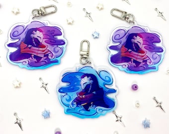 May include: Three acrylic keychains featuring a character design in shades of purple, blue, and red. Each keychain has a silver clasp. The background includes decorative stars, beads, and charms.