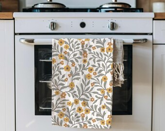 May include: A white kitchen towel with a floral pattern of yellow flowers and gray leaves. The towel is hanging on the oven door. A second, neutral-colored towel hangs next to it.