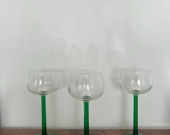 May include: Three clear glass wine glasses with green stems and bases. The glasses have a rounded bowl shape and are displayed on a wooden surface. The green stems and bases provide a striking contrast to the clear glass.