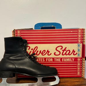 Może przedstawiać: Para czarnych skórzanych łyżew figurowych z srebrnymi ostrzami. Łyżwy znajdują się w oryginalnym pudełku, które jest czerwone w białe paski i z napisem "Silver Star Skates for the Family".