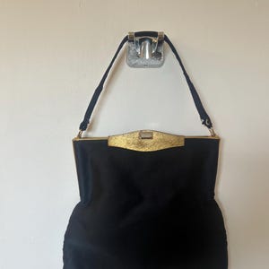 Può includere: Una borsa nera con chiusura e finiture color oro. La borsa ha una forma rettangolare con un manico. La borsa è appesa a un muro.