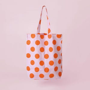 Könnte beinhalten: Eine rechteckige Tragetasche mit hellrosa Grundfarbe und einem sich wiederholenden Muster aus einfarbigen orangefarbenen Punkten. Die Tasche verfügt über zwei passende Stoffschulterriemen an der Oberkante, die ein einheitliches Design bieten.
