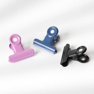 Könnte beinhalten: Drei Metall-Binderklammern in verschiedenen Farben: lila, blau und schwarz. Die Klammern sind zum Zusammenhalten von Papieren gedacht.