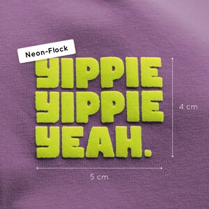 Pu&ograve; includere: Una superficie in tessuto viola con una grafica floccata al neon con il testo YIPPIE YIPPIE YEAH. La grafica misura 4 cm in altezza e 5 cm in larghezza, come indicato dalle linee di misurazione e dalle etichette visibili nell'immagine.