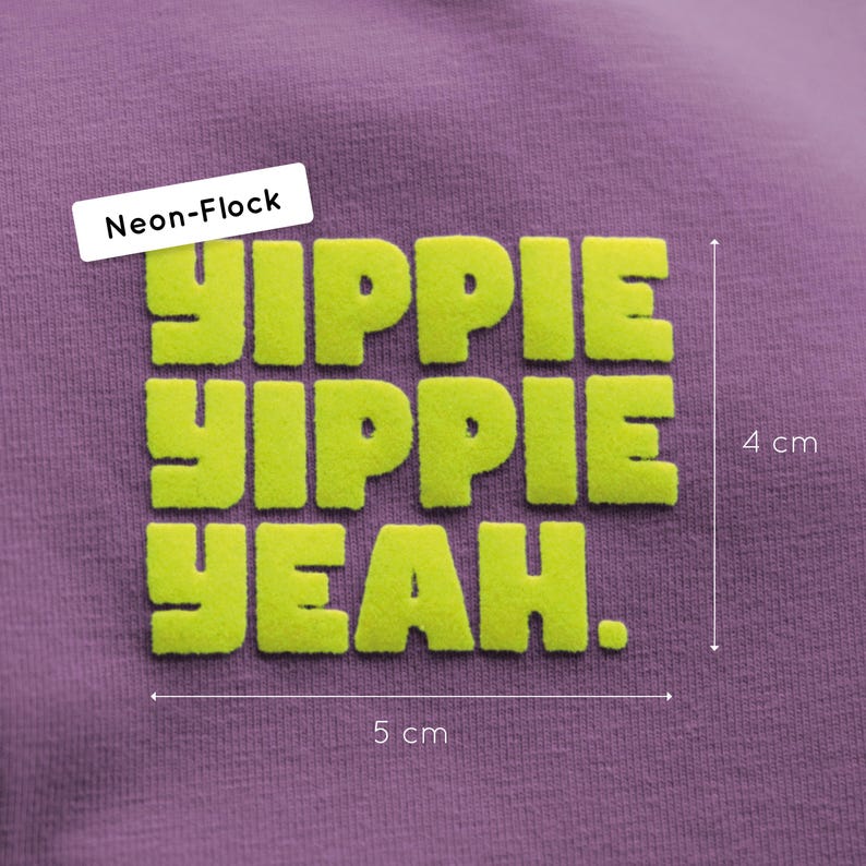Pu&ograve; includere: Una superficie in tessuto viola con una grafica floccata al neon con il testo YIPPIE YIPPIE YEAH. La grafica misura 4 cm in altezza e 5 cm in larghezza, come indicato dalle linee di misurazione e dalle etichette visibili nell'immagine.