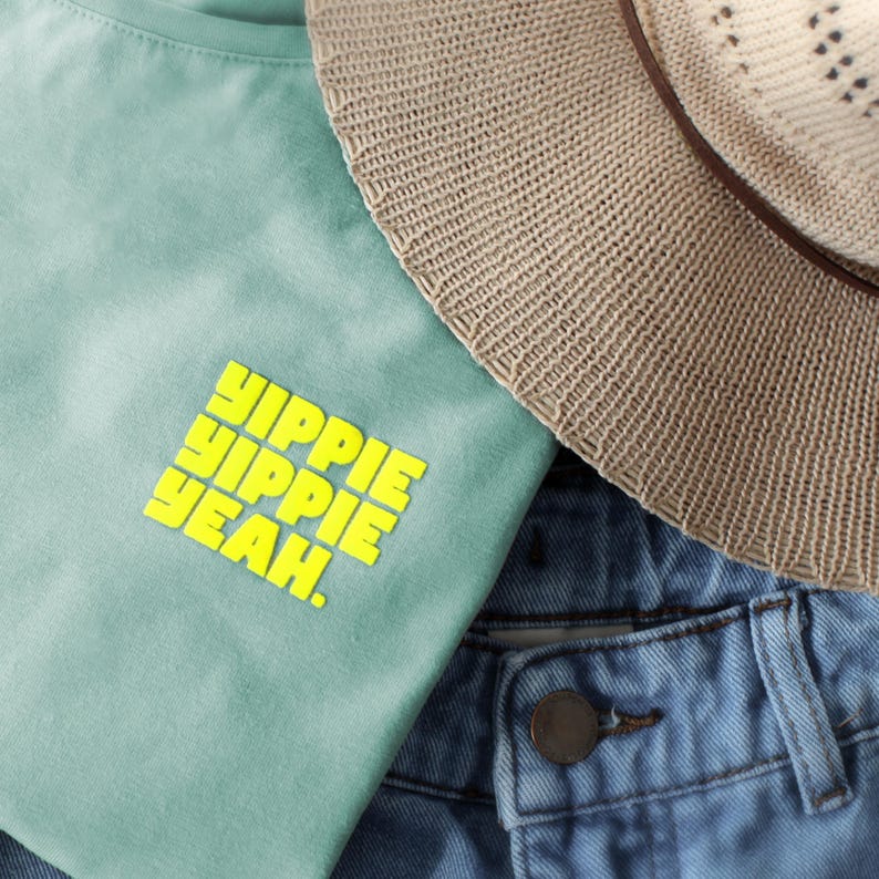 Pu&ograve; includere: Una t-shirt verde chiaro con il testo giallo brillante YIPPIE YIPPIE YEAH. stampato sul davanti, posizionata accanto a un cappello di paglia beige con una fascia marrone e un paio di jeans blu con un bottone metallico visibile in vita.