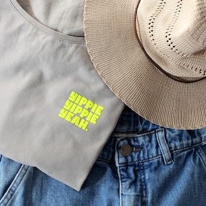 Könnte beinhalten: Ein lässiges Outfit mit einem hellgrauen T-Shirt mit dem Text "HIPPIE HIPPIE YEAH" in Neongrün, kombiniert mit blauen Jeans und einem beigen Strohhut mit braunem Band und Lochmuster.