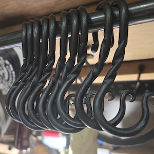 May include: A row of black, hand-forged metal S-hooks hanging from a metal rod. The hooks have a twisted design and are evenly spaced. The background is a wooden surface and a blurred workshop setting.