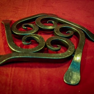 May include: A decorative, wrought iron trivet with a dark, metallic finish. The trivet features a stylized, swirling design with curved elements and a flat base. The piece is displayed on a red surface.