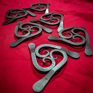 May include: A set of seven metal heart-shaped hooks with a swirling design. The hooks are arranged in a random pattern on a red surface.