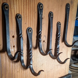 May include: Six black metal hooks with a twisted design are mounted on a wooden board. The hooks are all the same size and shape, and they have a small brass ring at the top. The board is made of light wood with a natural finish.