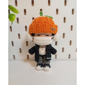 May include: A handmade crochet doll wearing a pumpkin-shaped hat, a white face mask, a black jacket, and gray pants. The doll has white ear coverings and black and white shoes. The doll is standing in front of a white background with evenly spaced holes.