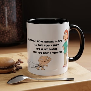 May include: White ceramic coffee mug with a black rim and handle. The mug features a cartoon image of a baby with a nappy and a woman. The text on the mug reads: "Mother, I come bearing a gift. I'll give you a hint: It's in my nappy, and it's not a toaster."