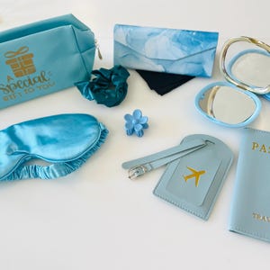 May include: A collection of light blue travel accessories, including a cosmetic bag with gold text, a sleep mask, a passport holder, a luggage tag, a scrunchie, a hair clip, a glasses case, a compact mirror, and a pair of socks.