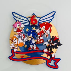 May include: A colorful cake topper featuring Sonic the Hedgehog and friends. The design includes Sonic, Tails, Amy, Knuckles, and Shadow, with a gold and blue background. The number 6 is prominently displayed, with a red and blue ribbon.