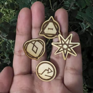 May include: Four small, wooden, laser-cut charms in various shapes: a triangle with a letter I, a heart with a diamond, a starburst, and a circle with an arrow. The charms are light brown with darker brown etched details.