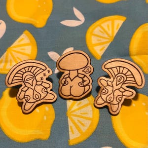 Peut inclure: Trois épingles en bois avec des dessins de contours noirs de figures en forme de champignon. L'une dort, une autre est en colère et la troisième a un design unique. Le fond est un tissu bleu avec des tranches de citron jaunes.